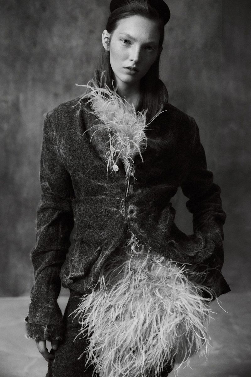 Monochrome fashion editorial portrait of a model wearing a textured jacket with feather accents and a headpiece against a textured wall.