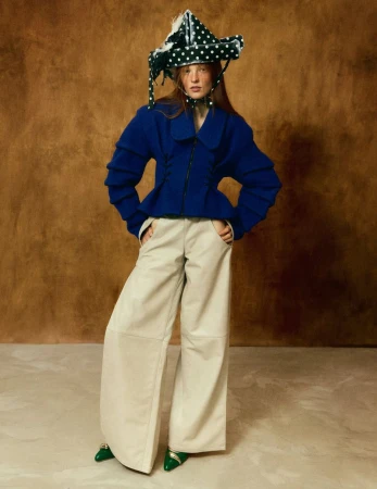 Model wearing a structured blue jacket, wide cream trousers, and a polka-dot hat against a textured brown background.