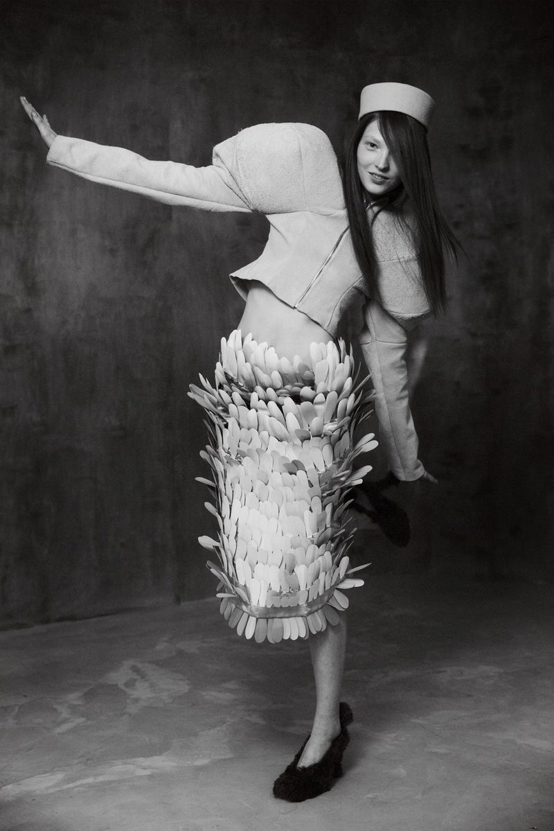 Model Zuza Bryk in structured jacket and feathered skirt striking a dynamic pose against a textured dark backdrop for Elle Hungary.