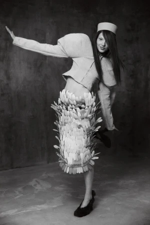 Model Zuza Bryk in structured jacket and feathered skirt striking a dynamic pose against a textured dark backdrop for Elle Hungary.