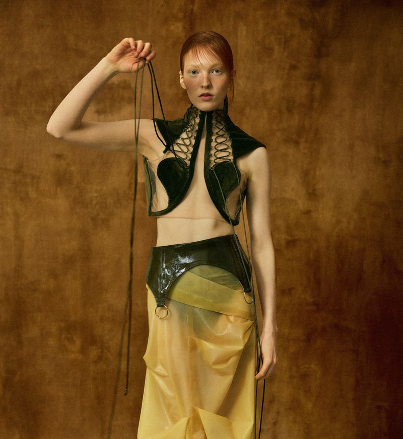 Red-haired model posing in a green lace-up vest and translucent yellow skirt against a warm textured brown backdrop