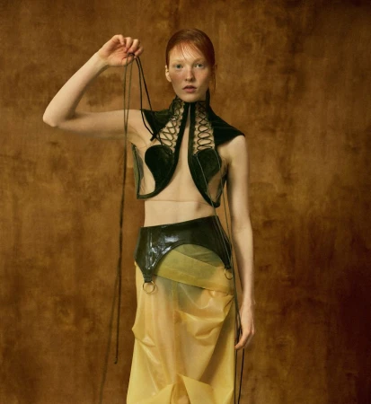 Red-haired model posing in a green lace-up vest and translucent yellow skirt against a warm textured brown backdrop