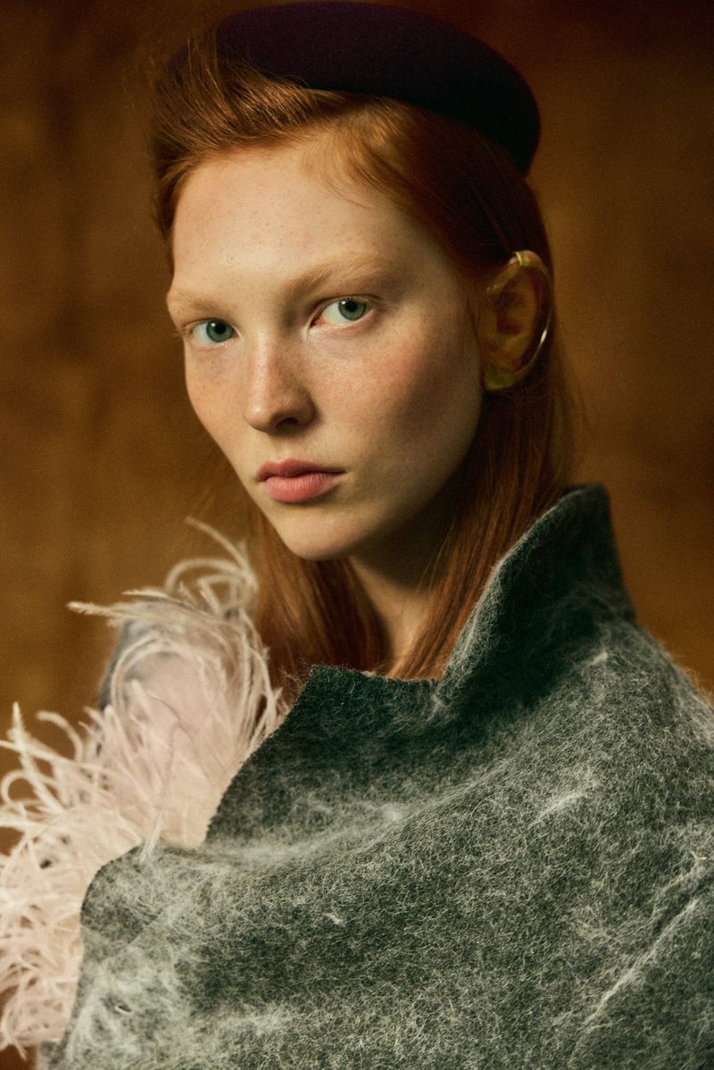 Red-haired model wearing a textured grey coat with feather trim and a dark hat in a portrait for Elle Hungary