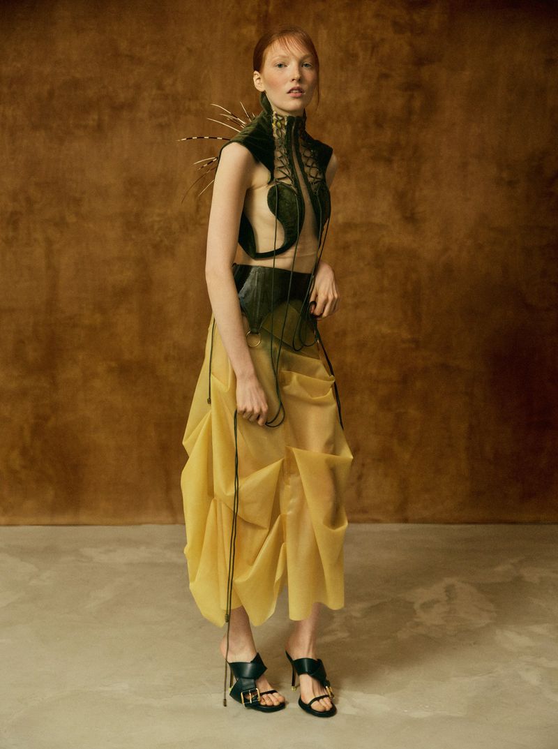 Model wearing a structured dark green top and translucent yellow skirt against a warm textured backdrop.