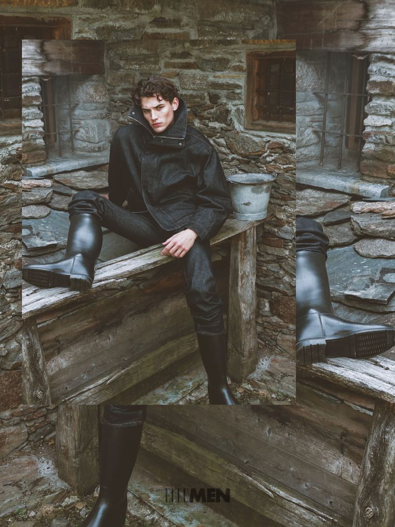 Male model in a dark jacket and high black rubber boots seated outdoors against a rustic stone wall
