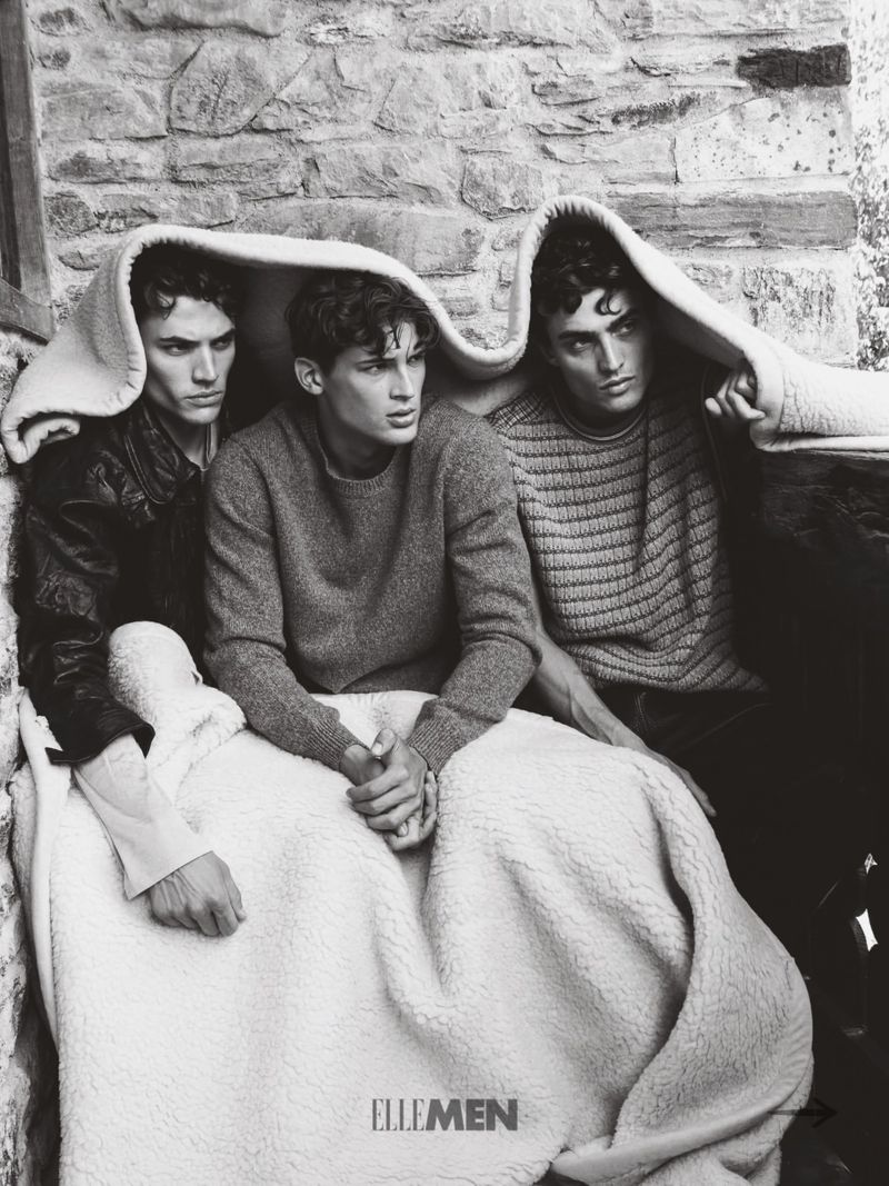 Three male models wrapped in a large white sherpa blanket against a rustic stone wall background