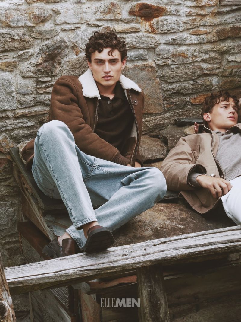 Two male models seated outdoors against a rough stone wall wearing winter jackets and denim jeans