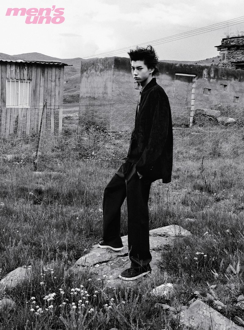 Black and white editorial portrait of a young male model standing on a rocky path in front of rustic buildings.