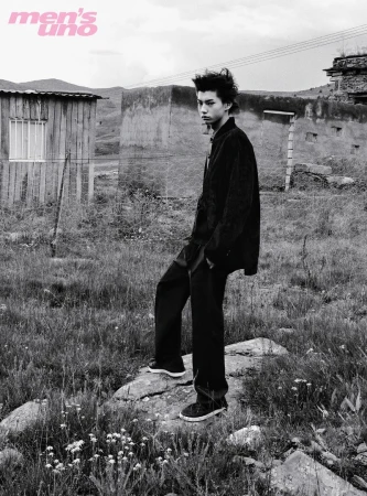 Black and white editorial portrait of a young male model standing on a rocky path in front of rustic buildings.
