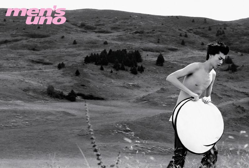 A shirtless male model holding a large circular reflector while walking across a grassy hill landscape.