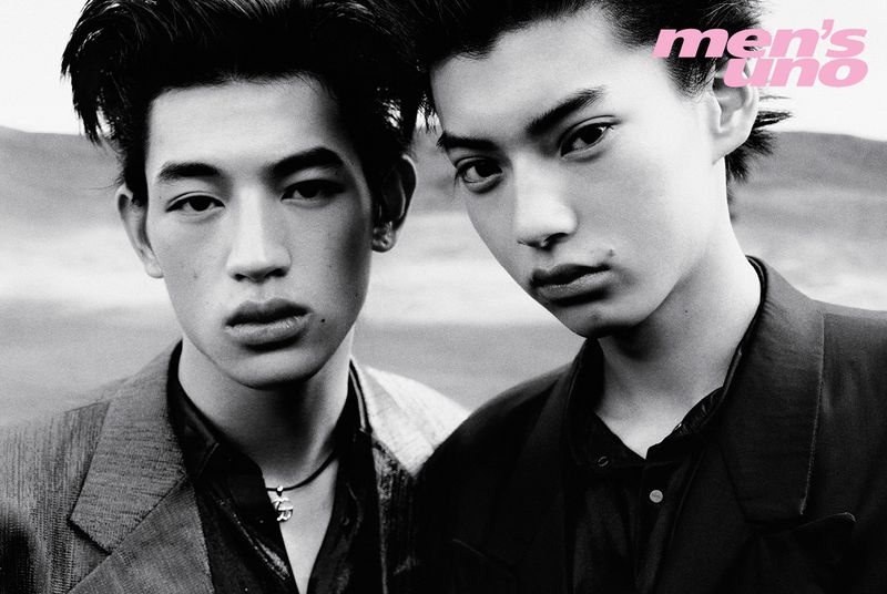 Black and white portrait of two male models with dark hair wearing dark tailored jackets against an open-air background.