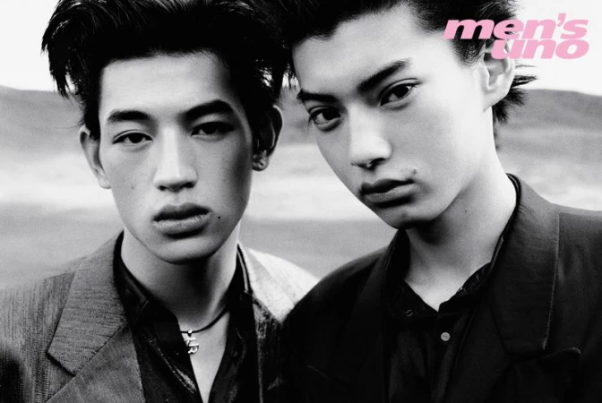 Black and white portrait of two male models with dark hair wearing dark tailored jackets against an open-air background.