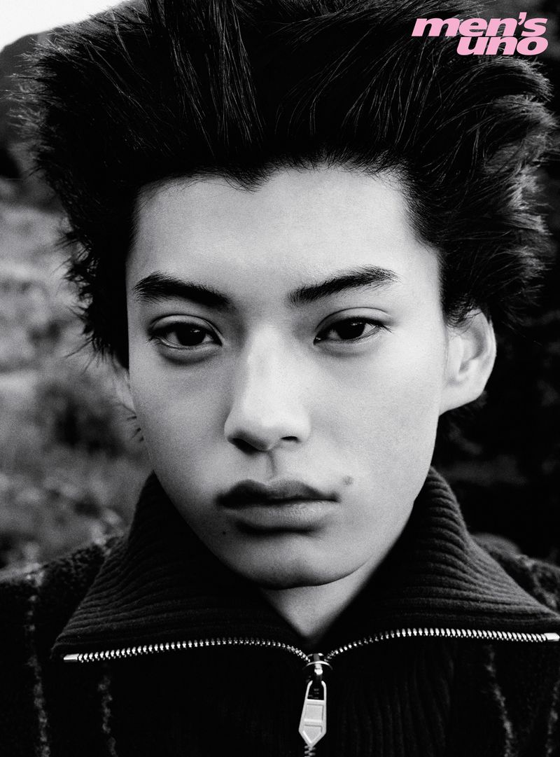 Close-up black and white portrait of a young male model with voluminous spiky hair wearing a zipped turtleneck top.