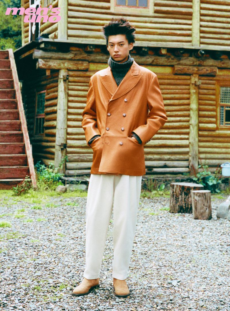 Model wearing a tan leather double-breasted jacket and white trousers standing outdoors before a wooden cabin.