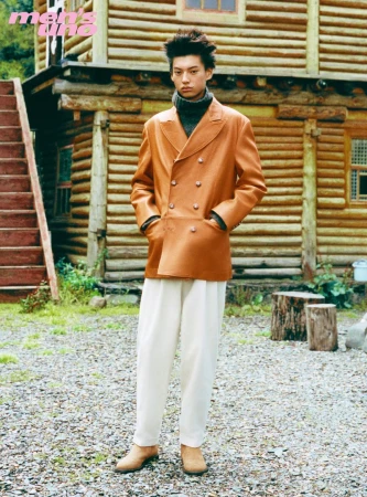 Model wearing a tan leather double-breasted jacket and white trousers standing outdoors before a wooden cabin.