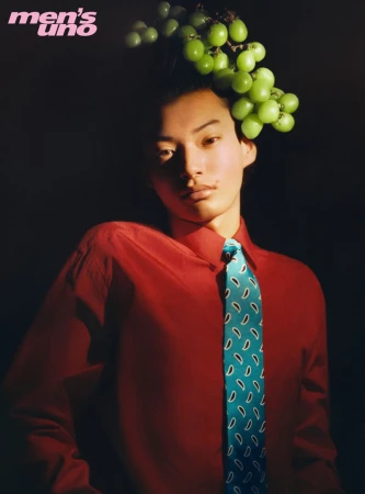 Model wearing a red shirt and turquoise patterned tie with green grapes styled in their hair against a dark background.