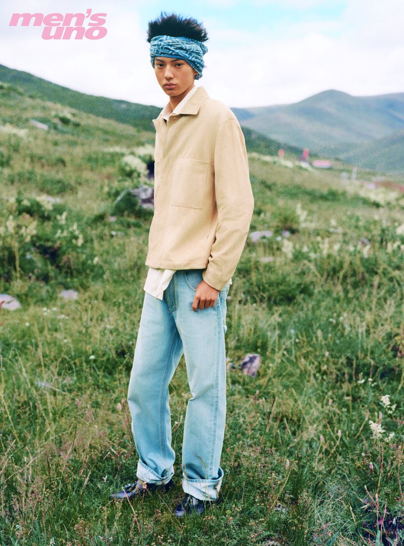 Model wearing a tan jacket and blue jeans standing in a lush green meadow for Men's Uno China