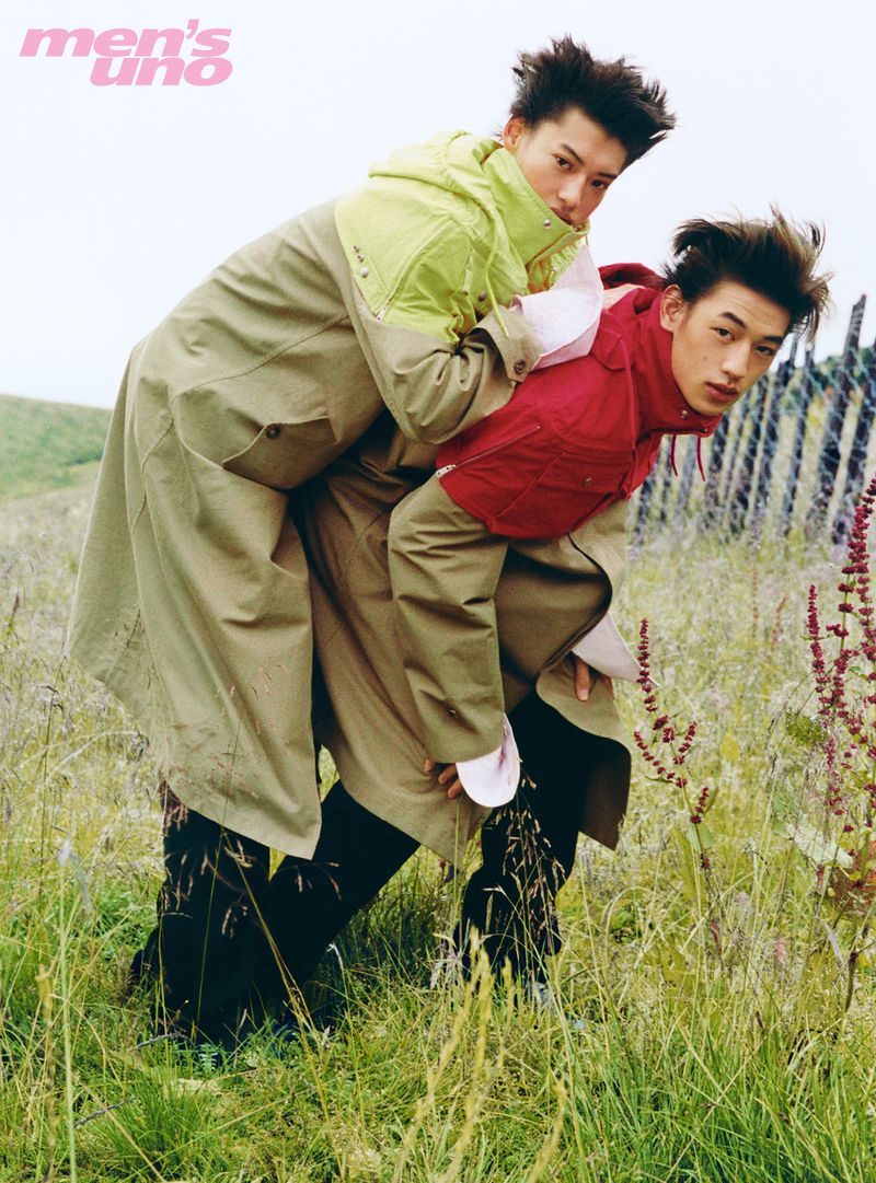 Two male models posing together in an outdoor meadow field wearing layered coats for a Men's Uno China fashion editorial.