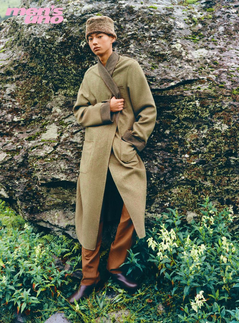Model wearing an olive coat and fur hat standing outdoors against a large rock formation