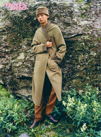 Model wearing an olive coat and fur hat standing outdoors against a large rock formation