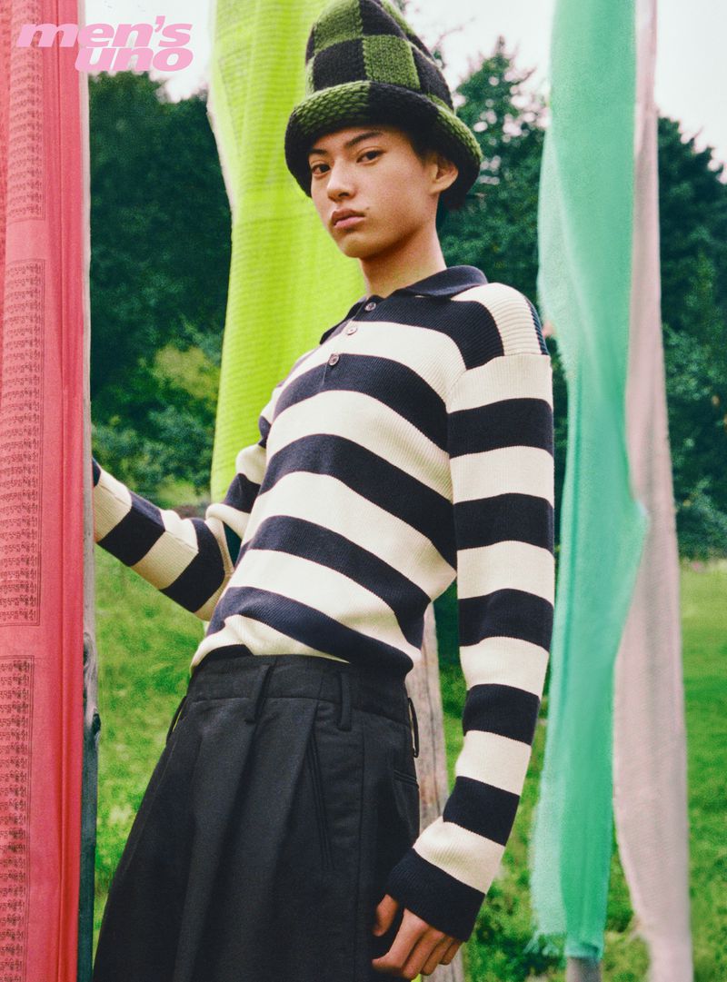 Model wearing a black and white striped polo shirt, black trousers, and a green checked hat outdoors among vertical fabric banners
