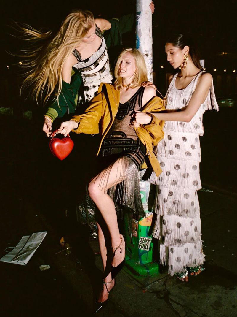 Three women in fashionable dresses pose in an urban setting at night. One holds a red heart-shaped purse, and street features visible.