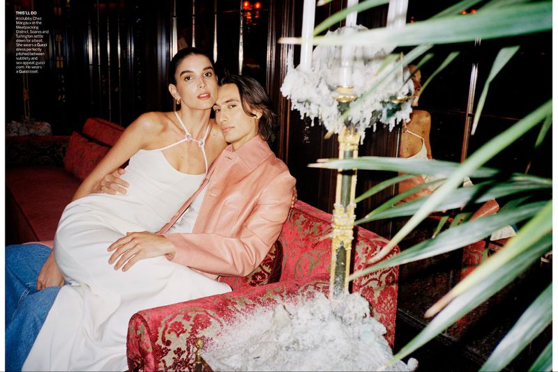 Stylish couple poses indoors, the woman in a white dress and the man in a pink jacket, seated on ornate seating amidst green foliage.