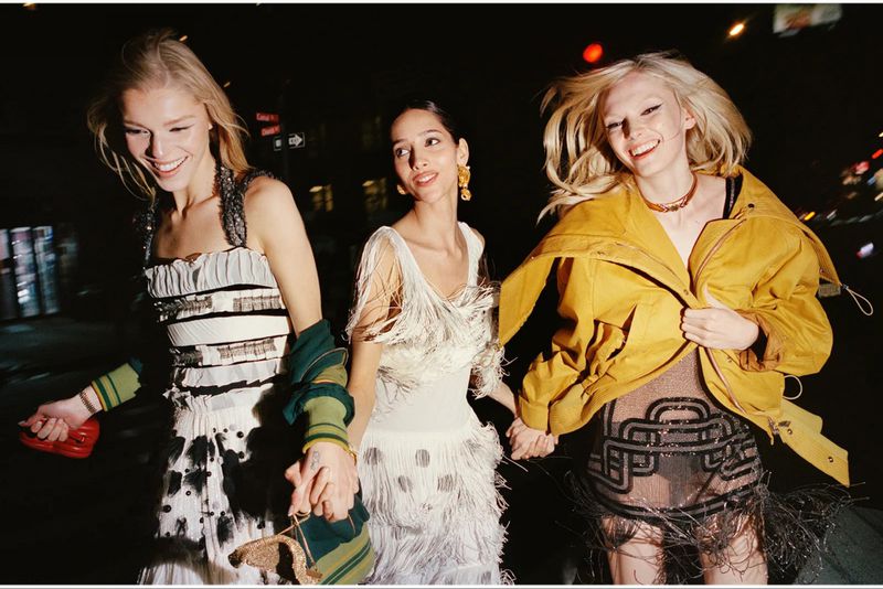Three smiling women in dresses hold hands on a dark city street at night, illuminated by streetlights and car lights.