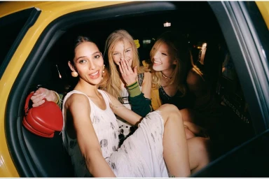 Three female models smiling inside the back of a yellow taxi cab holding a red heart-shaped handbag for American Vogue.