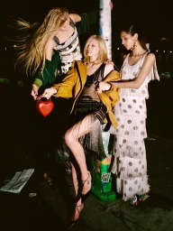 Three models in evening wear, including a fringed dress and tiered gown, pose against a street pole at night holding a red heart purse.