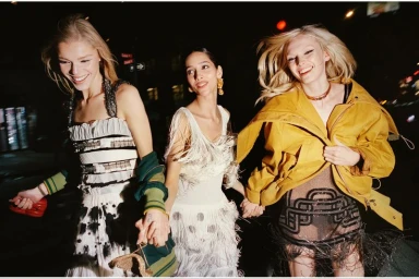 Three models holding hands and smiling while walking down a dark city street at night in fringed and textured designer eveningwear.