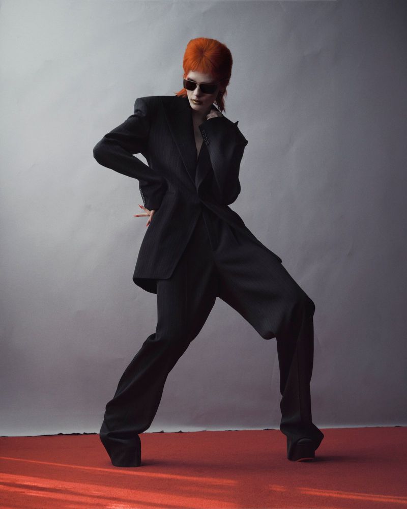 Model Leticia Hall with bright orange hair wearing a black oversized suit and sunglasses against a grey studio backdrop
