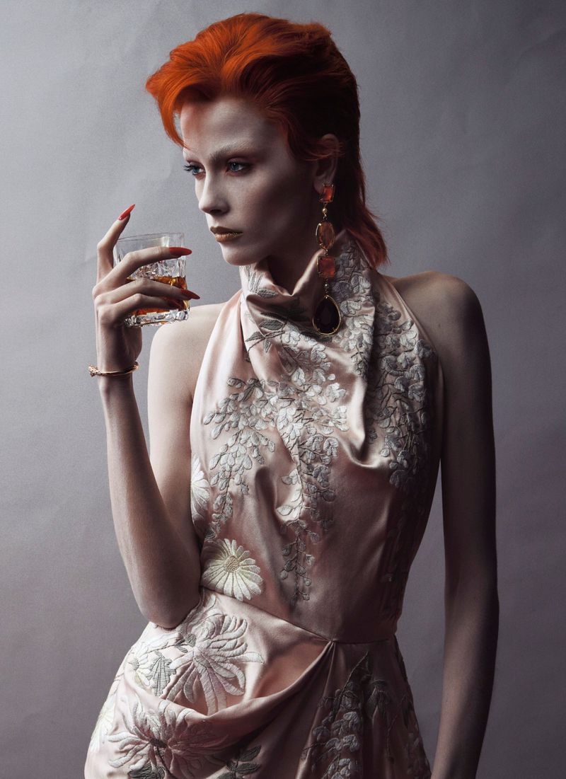 Model Leticia Hall with bright red hair wearing an embroidered halter dress holding a glass of whiskey for V Magazine