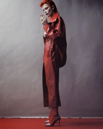 Model with red hair wearing a long red coat standing against a grey seamless studio backdrop.