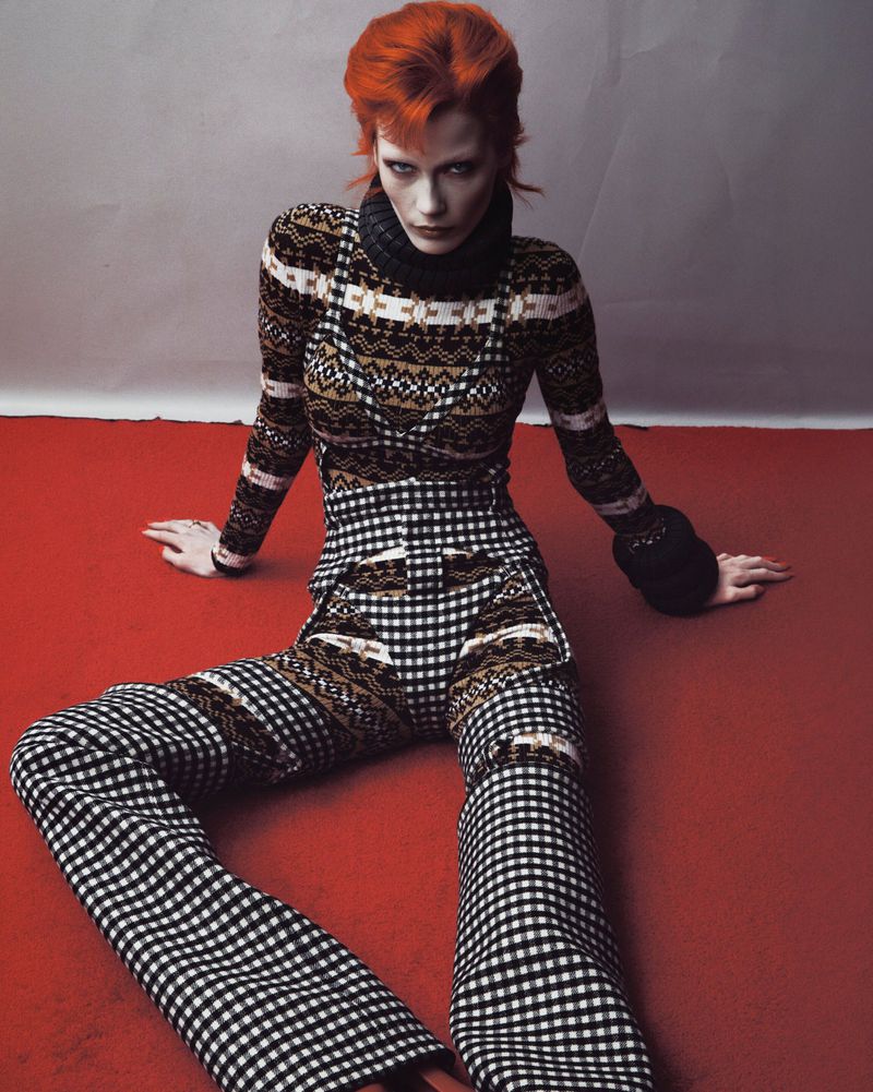 Model Leticia Hall with bright red hair wearing patterned knitwear and checkered pants seated on a red floor