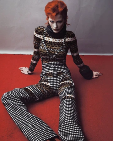 Model Leticia Hall with bright red hair wearing patterned knitwear and checkered pants seated on a red floor