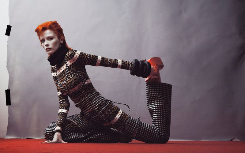 Model Leticia Hall with vibrant orange hair in patterned turtleneck and trousers stretching on a red floor against a grey backdrop