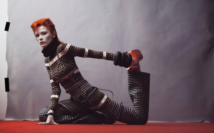 Model Leticia Hall with vibrant orange hair in patterned turtleneck and trousers stretching on a red floor against a grey backdrop