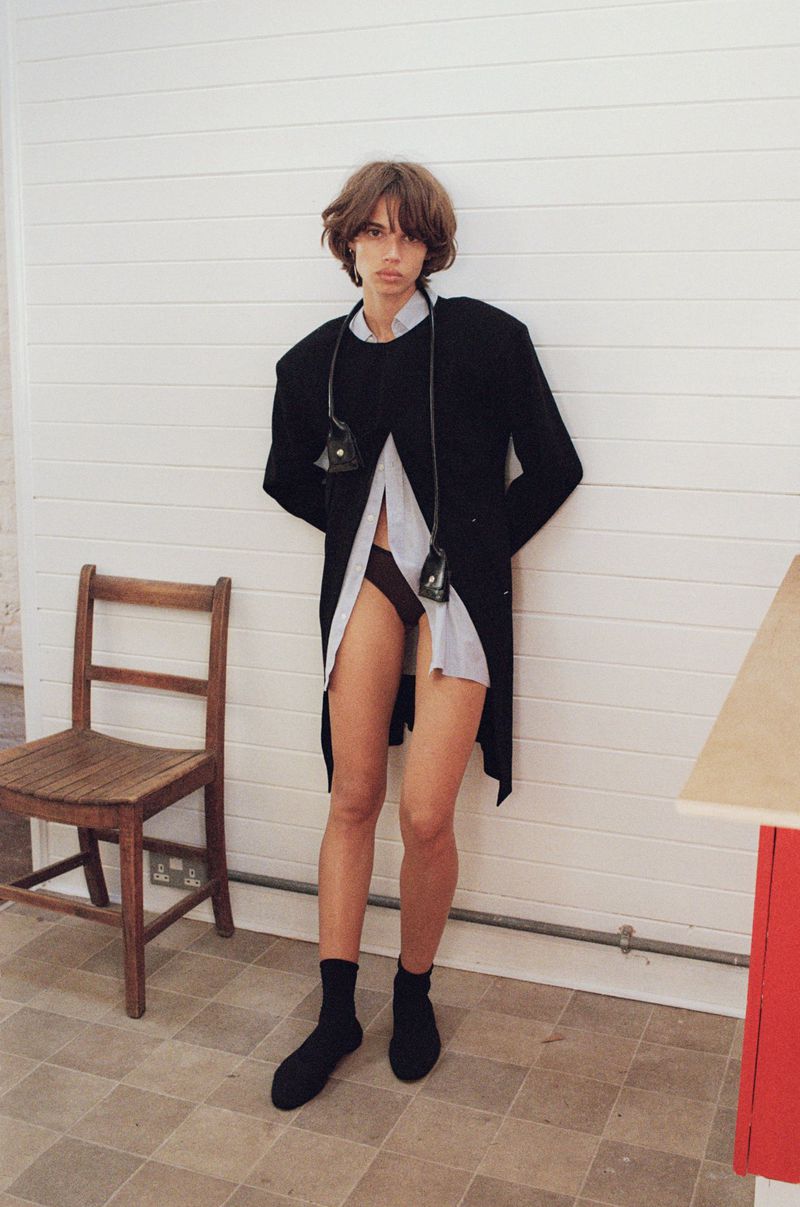 Model Mathilda Gvarliani wearing a black coat and buttoned shirt against a white panelled wall in a Pop Magazine editorial shot.