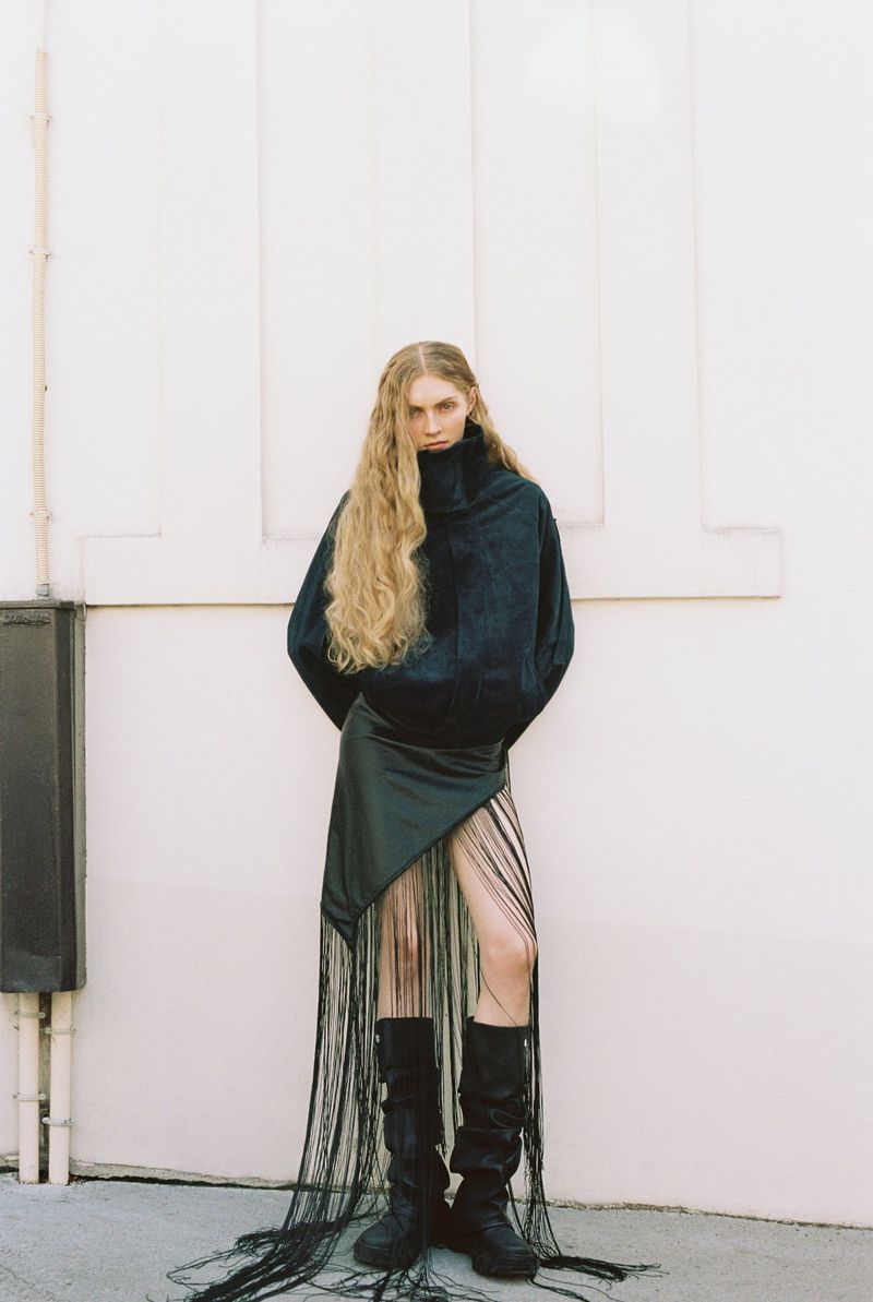 Model with long blonde hair wearing a black jacket and fringed skirt against a white wall
