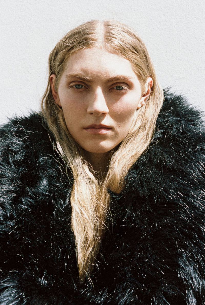 Close-up portrait of a model with long blonde hair wearing a black furry textured coat against a plain light wall.