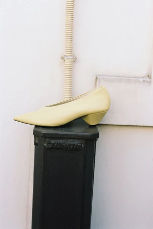 Pale yellow pointed-toe pump resting on a dark post against a light wall for ONE Magazine editorial.