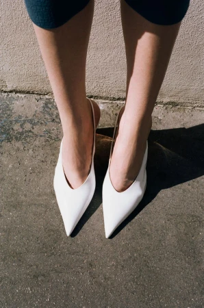 Close-up of a person's legs wearing pointed white slingback shoes standing against a textured concrete wall.