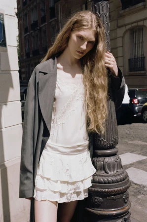 Model with long blonde hair leaning against a dark ornate lamppost on an urban street wearing a white slip dress and dark coat