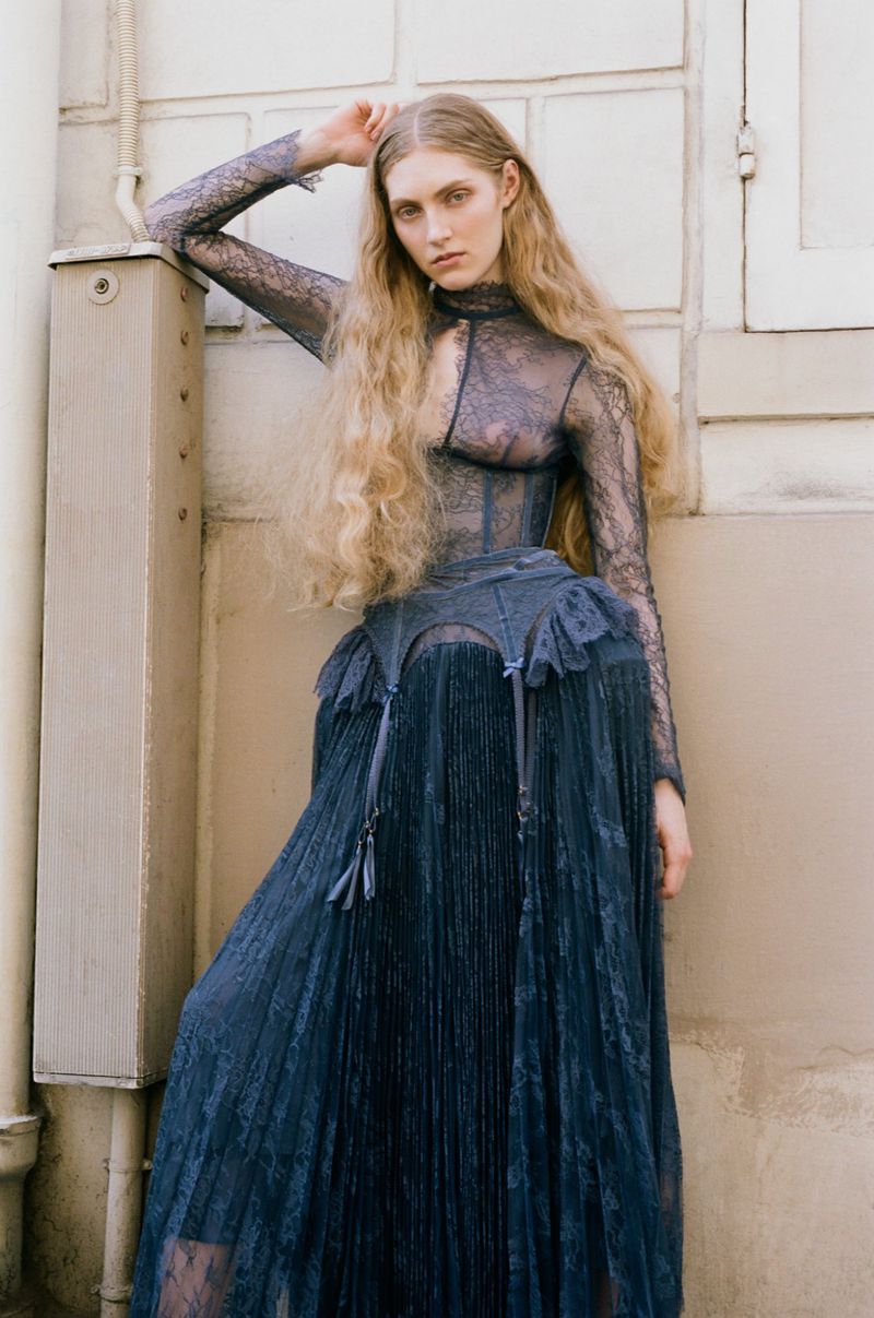 Model Constanze Van Rosmalen in a sheer blue lace dress leaning against a light architectural wall for ONE Magazine.