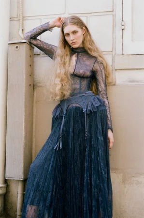 Model Constanze Van Rosmalen in a sheer blue lace dress leaning against a light architectural wall for ONE Magazine.