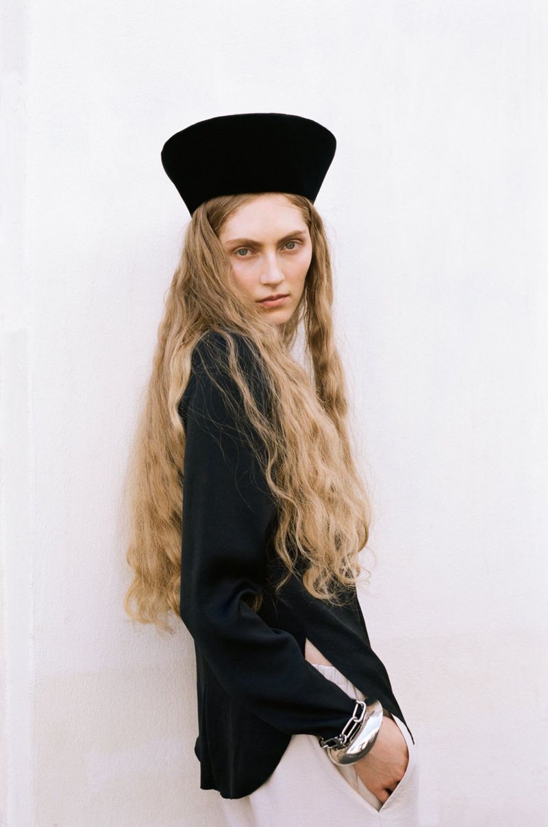 Model Constanze Van Rosmalen wearing a black pillbox hat and dark top against a white wall for ONE Magazine.
