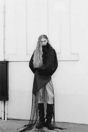Model with long wavy hair wearing a dark turtleneck and fringe skirt against an exterior wall