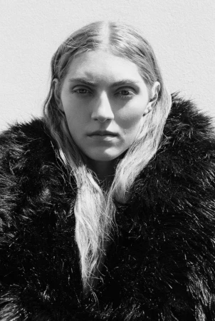 Portrait of a female model wearing a textured black faux fur coat against a light wall in monochrome.