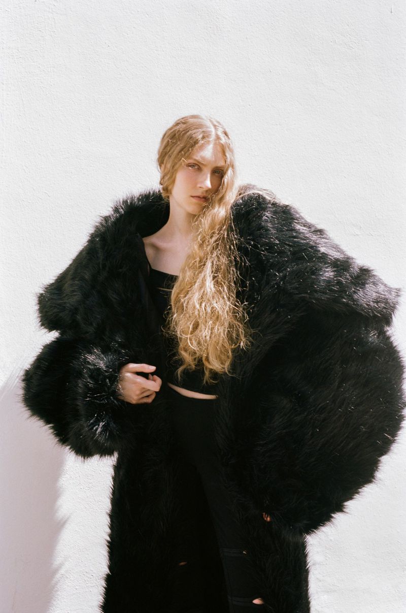 Model with long blonde hair wearing a voluminous black fur coat against a bright white wall